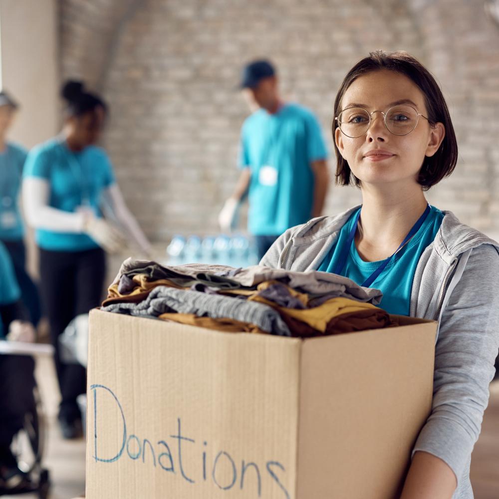 Young woman holding donation box full of clothes while volunteering at community center and looking at camera.