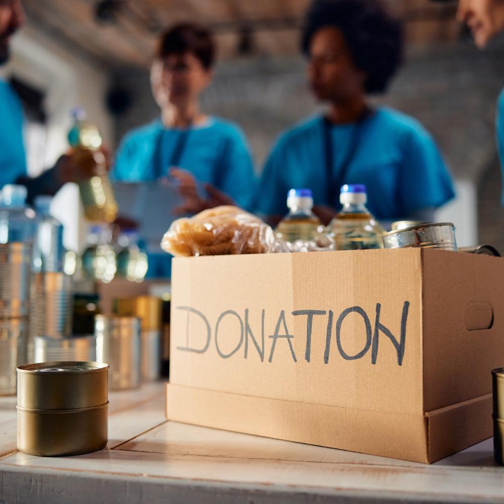 Cardboard box full of donated groceries at food bank with group of volunteers in the background.